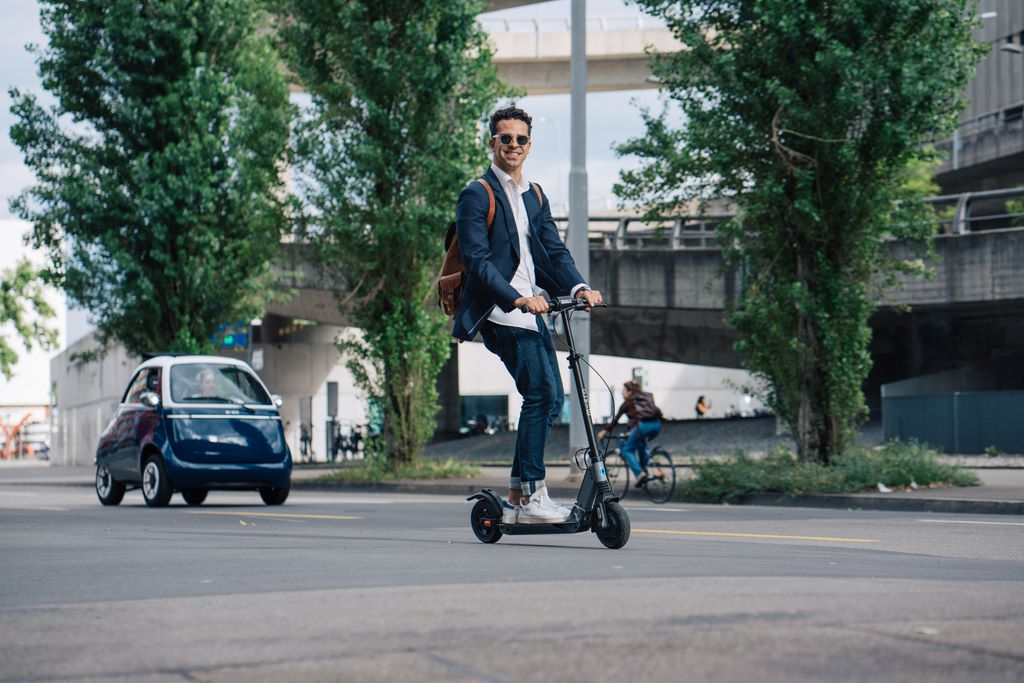 un homme roulant en trottinette electrique micro mobility merlin II en pleine ville suivi par une voiture micro lino